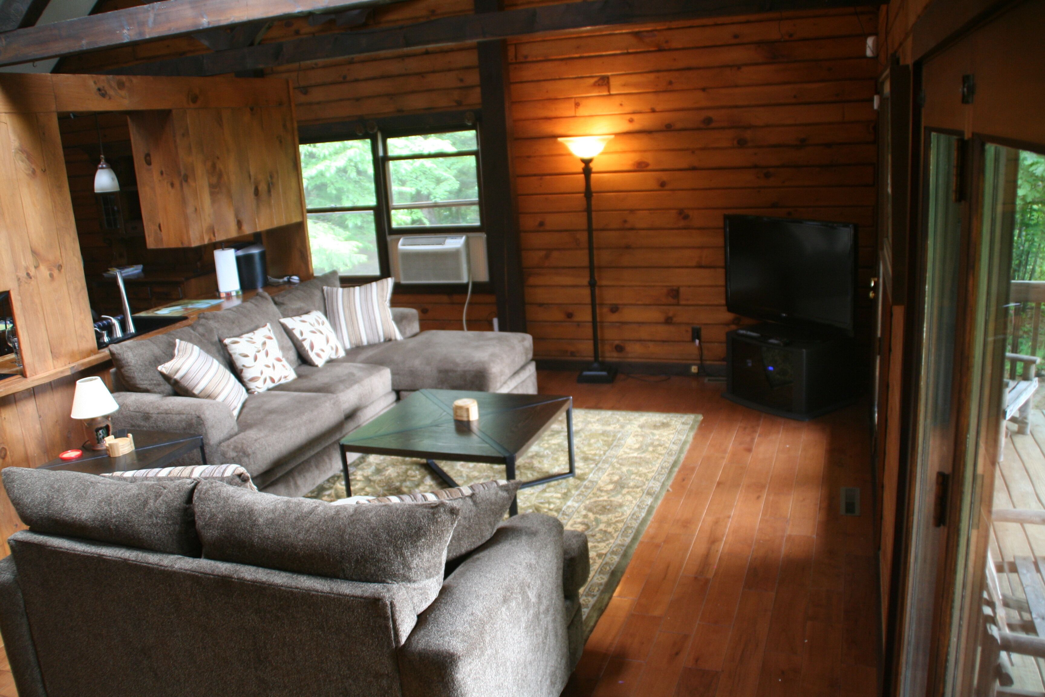 Un intérieur de chalet chaleureux avec un canapé d’angle, une petite table, un téléviseur et une grande fenêtre.