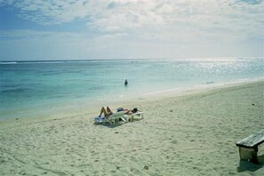 A beach with two people lying on sun loungers, a wooden bench, and a calm sea. - Aroa Beachfront House (Rarotonga)