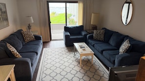 A living room with two dark sofas, a coffee table, a patterned rug, and a view of a green lawn and boats through a glass door.