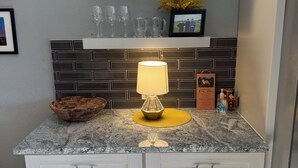 Una cocina con encimera de granito, armarios blancos, estante colgante con tazas y una foto enmarcada.