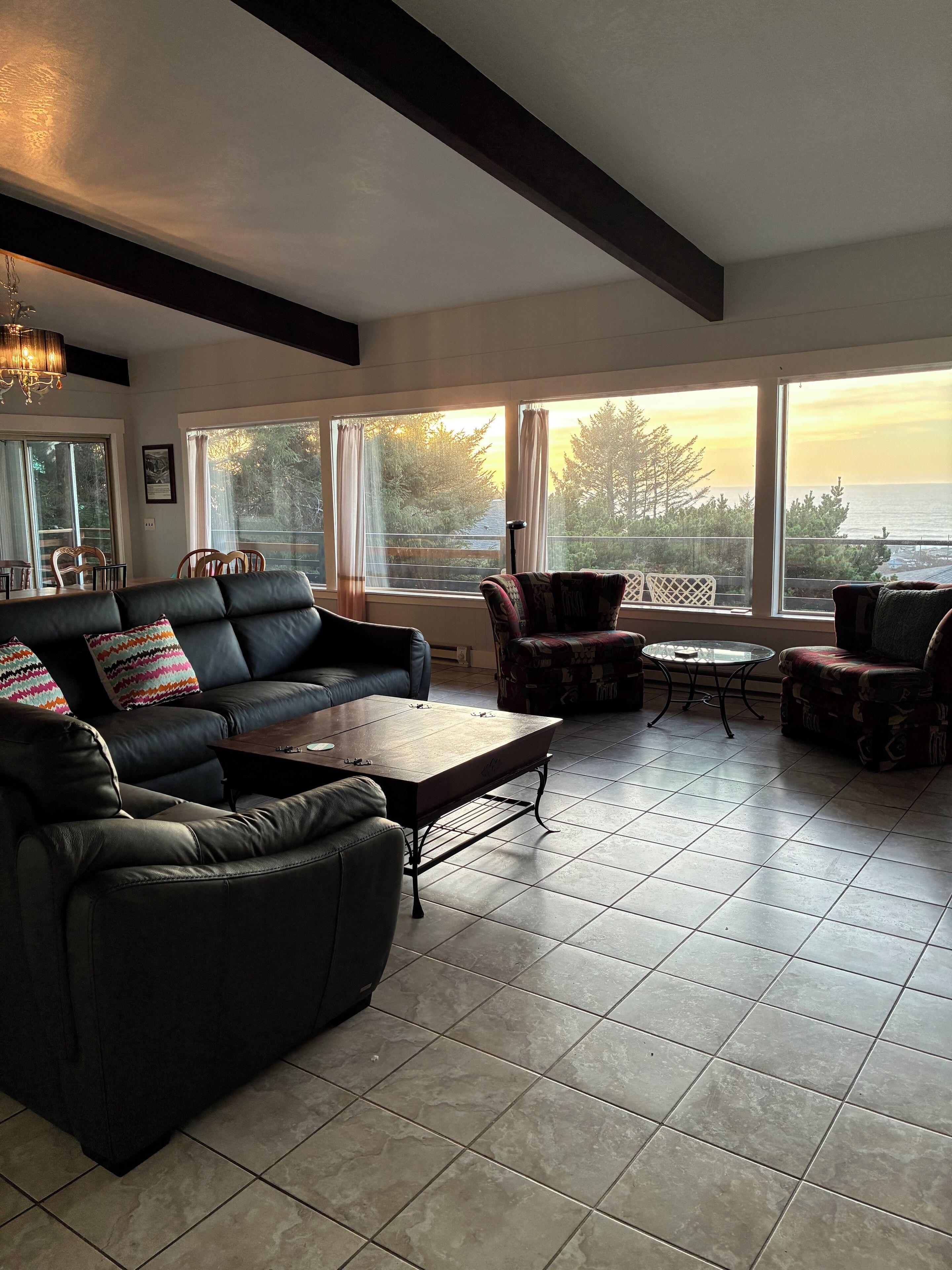 A modern living room with a leather sofa, patterned armchairs, a coffee table, and large windows offering a view of the sea.