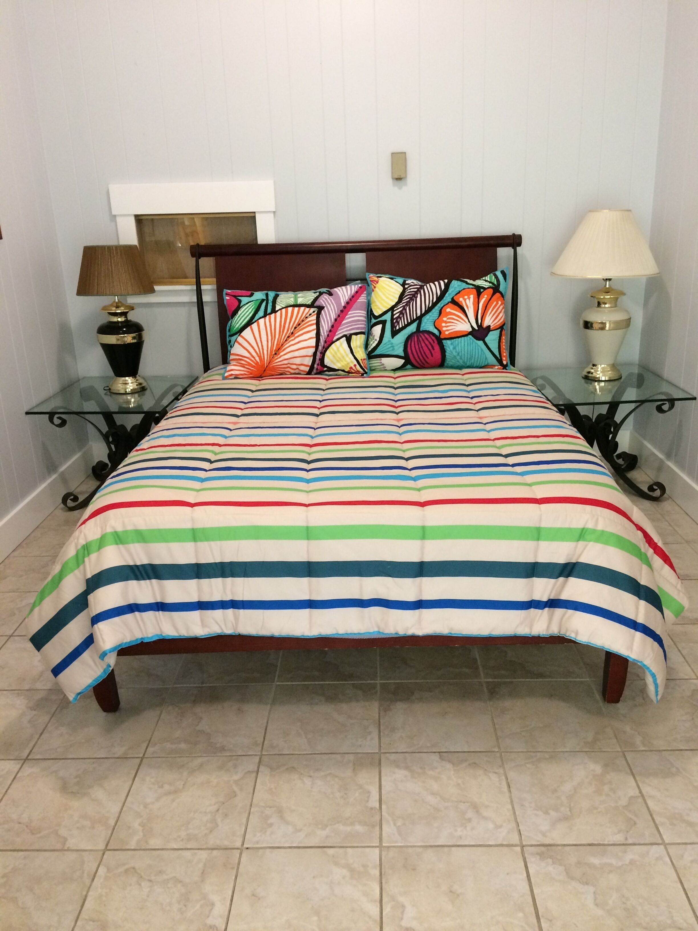 Una cama con colcha a rayas, dos mesitas de noche con lámparas y un cabecero con almohadones decorativos.