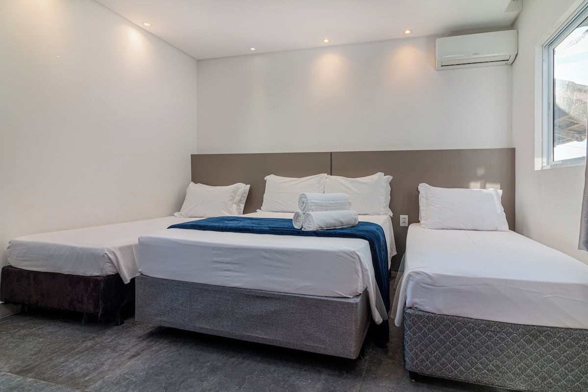 A hotel room with three beds, a large window, and an air conditioning unit.