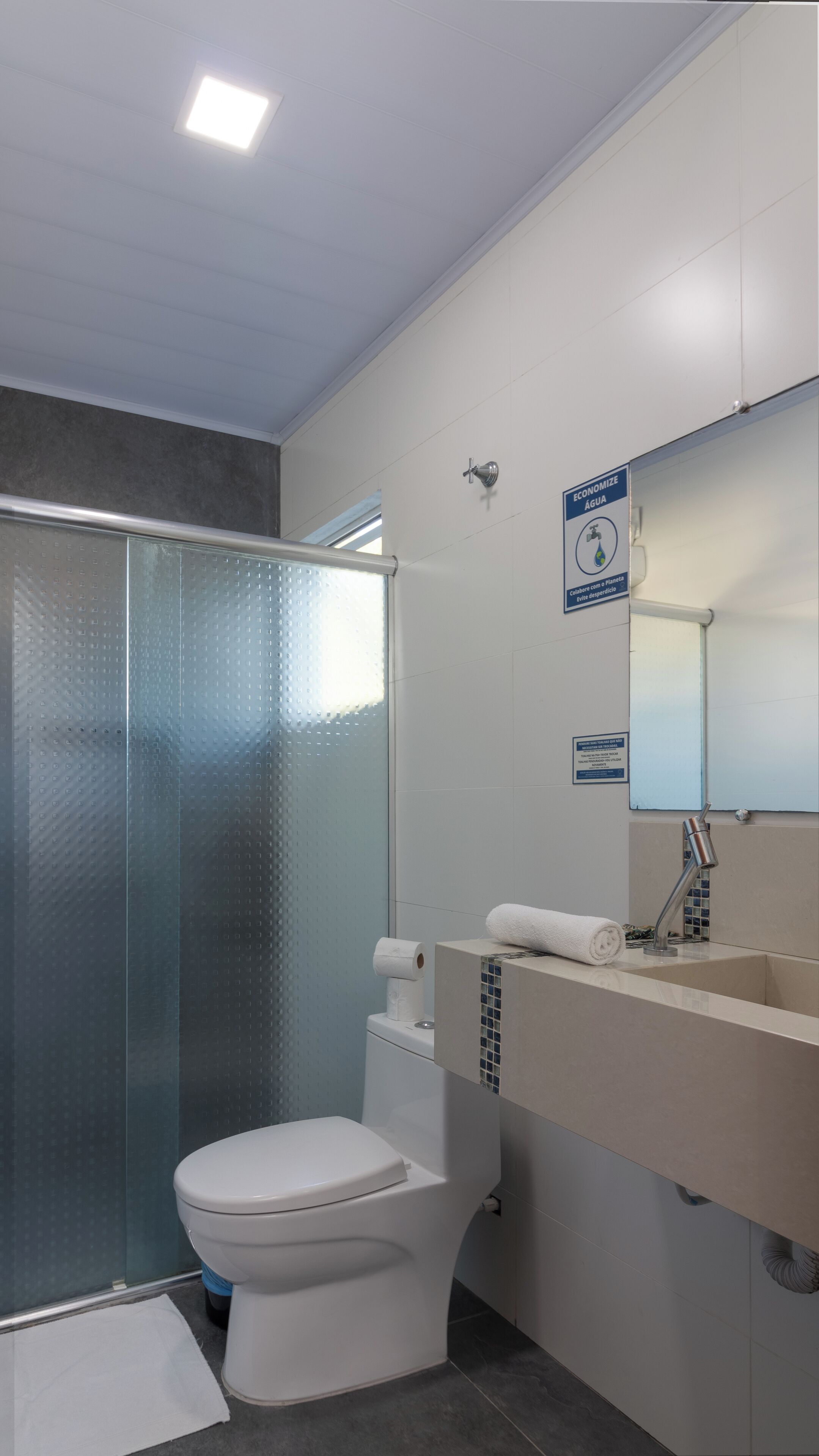 Egy modern fürőszoba, amely tartalmaz egy WC-t, egy üvegfalas zuhanykabint, egy mosdókagylót tükörrel, és egy törölközőtartót.