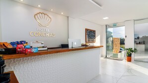A hotel reception area with a white textured wall, a wooden reception desk, and a large window offering a view of the outdoors. - Hotel Conchas (Penha)