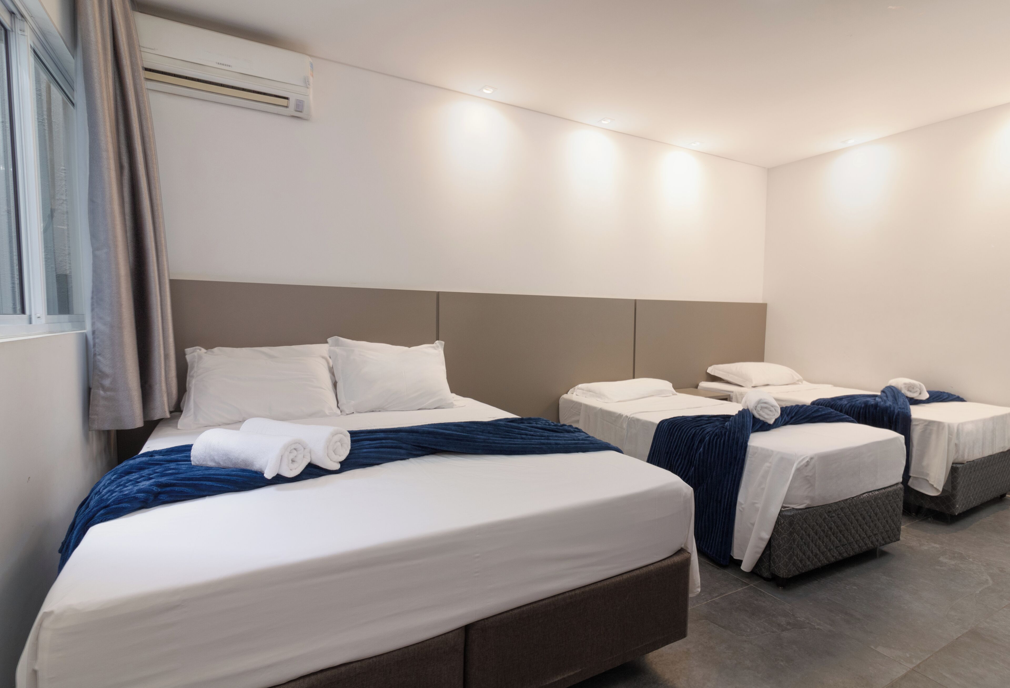 Una habitación de hotel con una cama grande, dos camas más pequeñas, un cabecero y una ventana con cortinas.