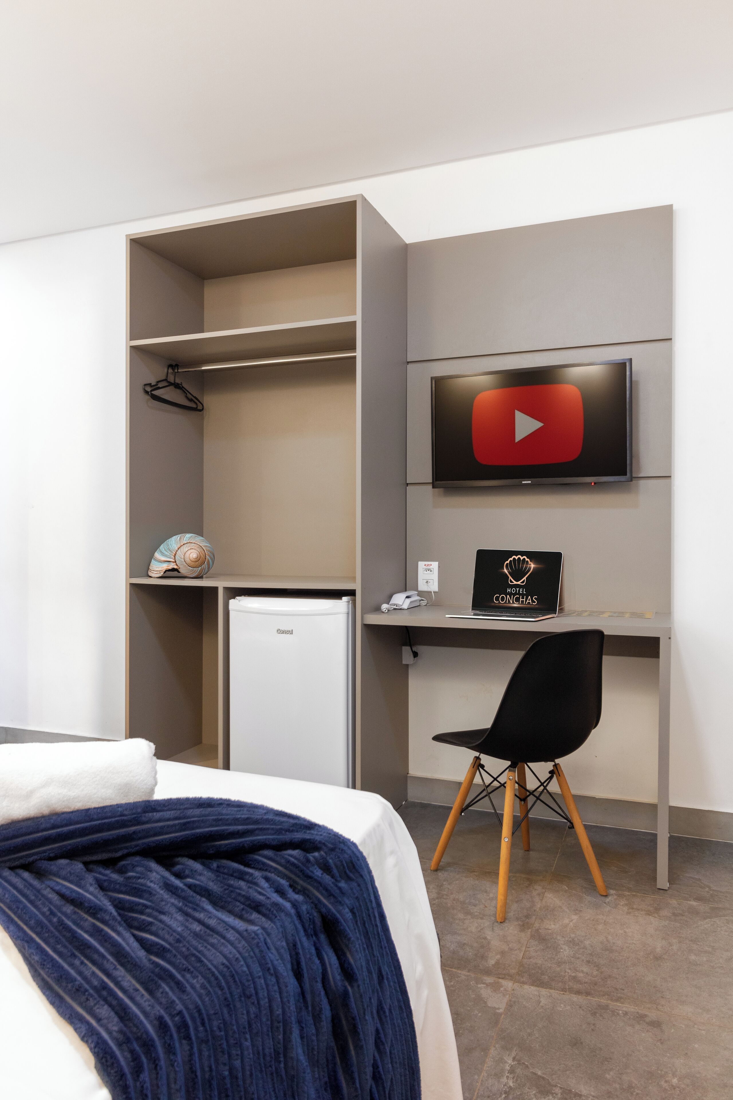 A hotel room with a bed, a desk with a laptop, a TV, and a small refrigerator.
