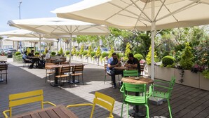 An outdoor café with wooden tables and chairs, large white umbrellas, and lush greenery in the background. - The Spot Hostel (Tel Aviv)