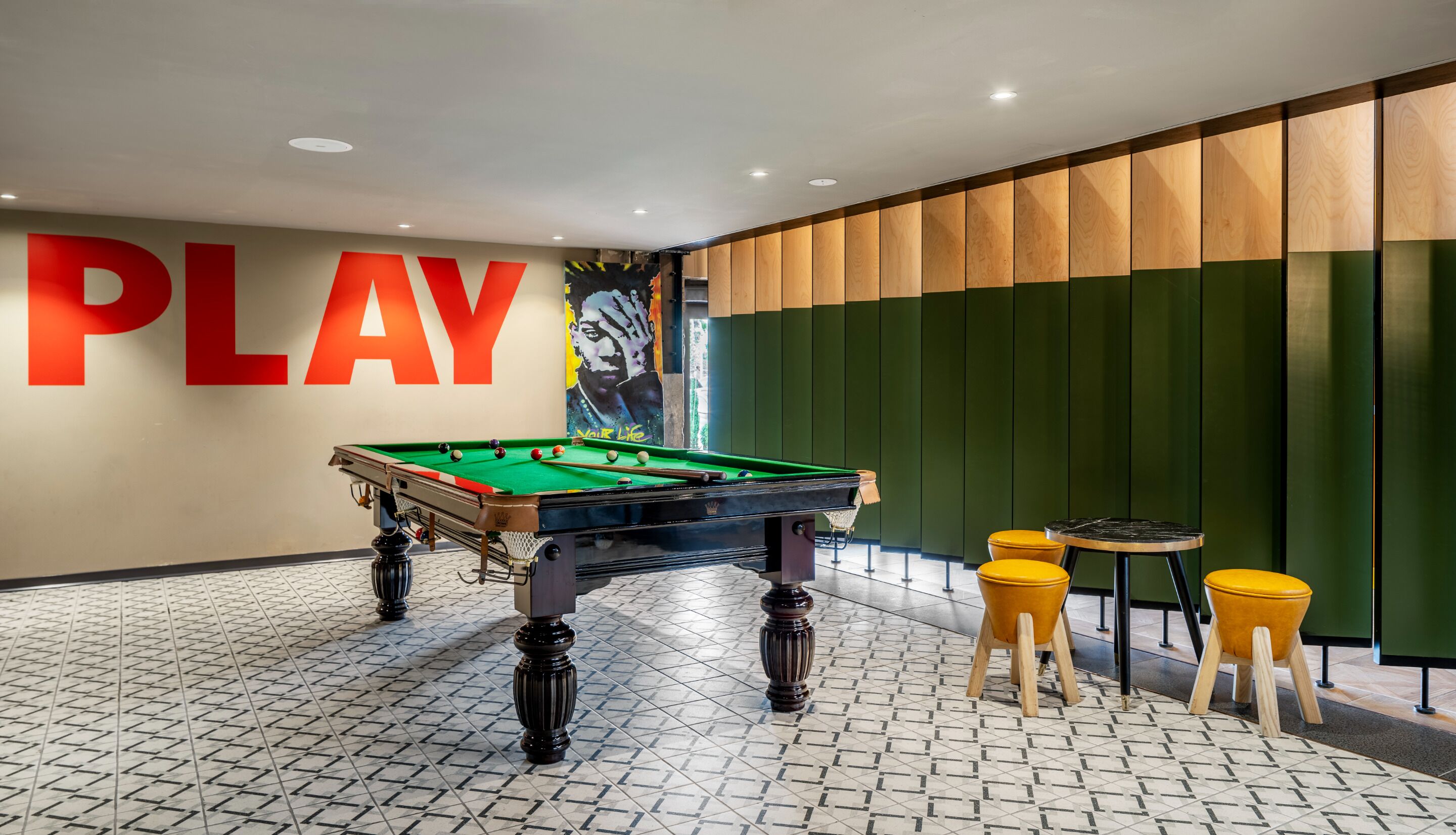 A pool table in a room with a 'PLAY' sign, green and wooden panel walls, and yellow stools.