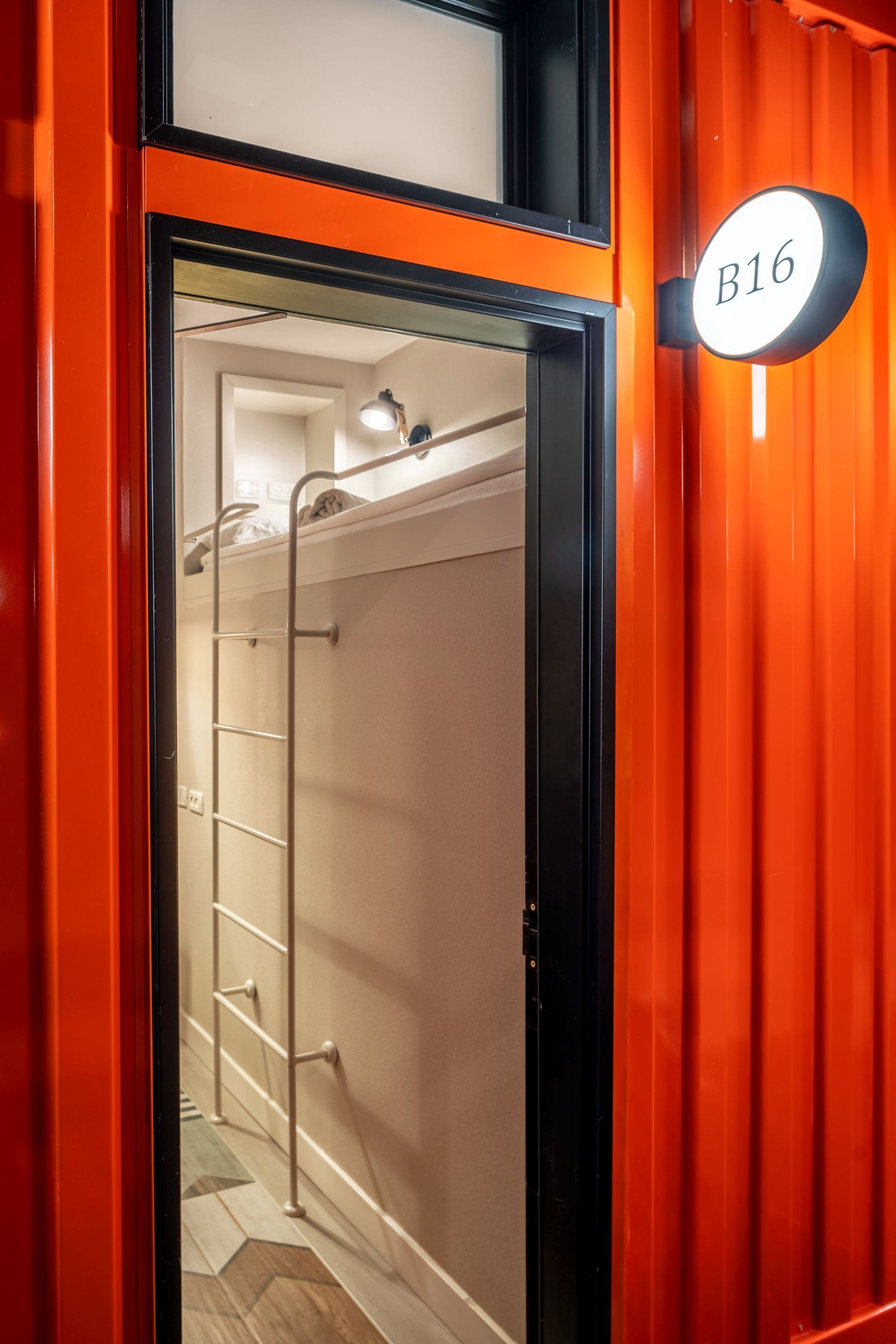 A corridor with a metal ladder leading to a room with a bunk bed. A sign with "B16" is visible on the wall.