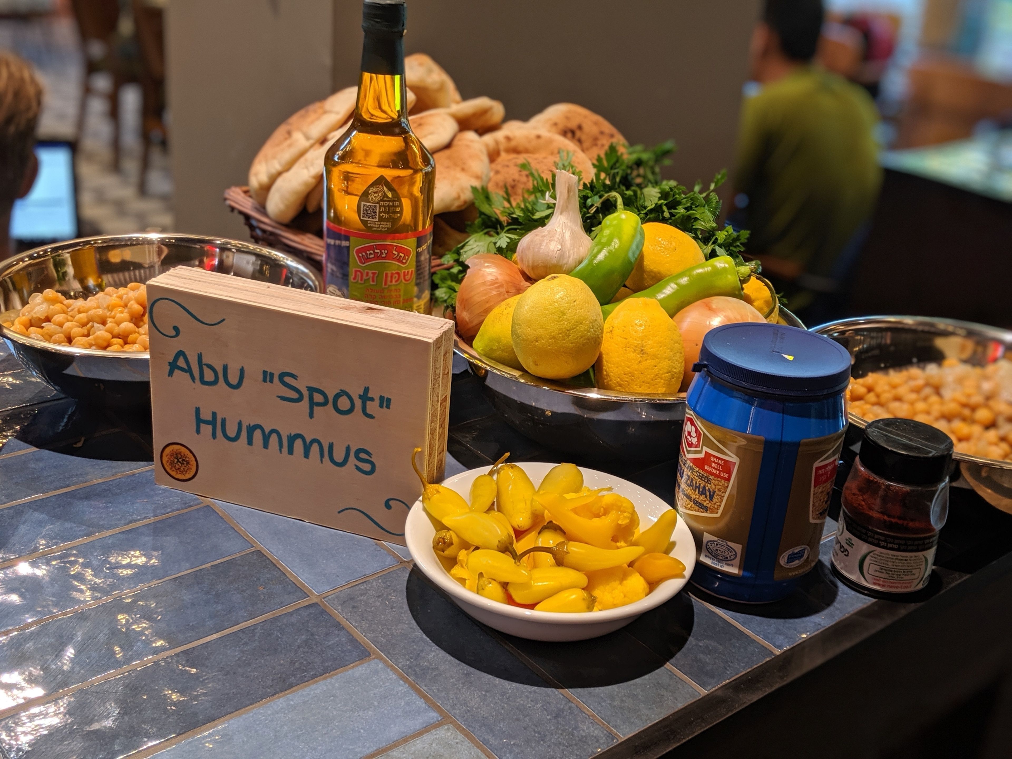 Una mesa de buffet con diversos platos y aderezos, incluyendo hummus, pan, frutas y una botella de aceite.
