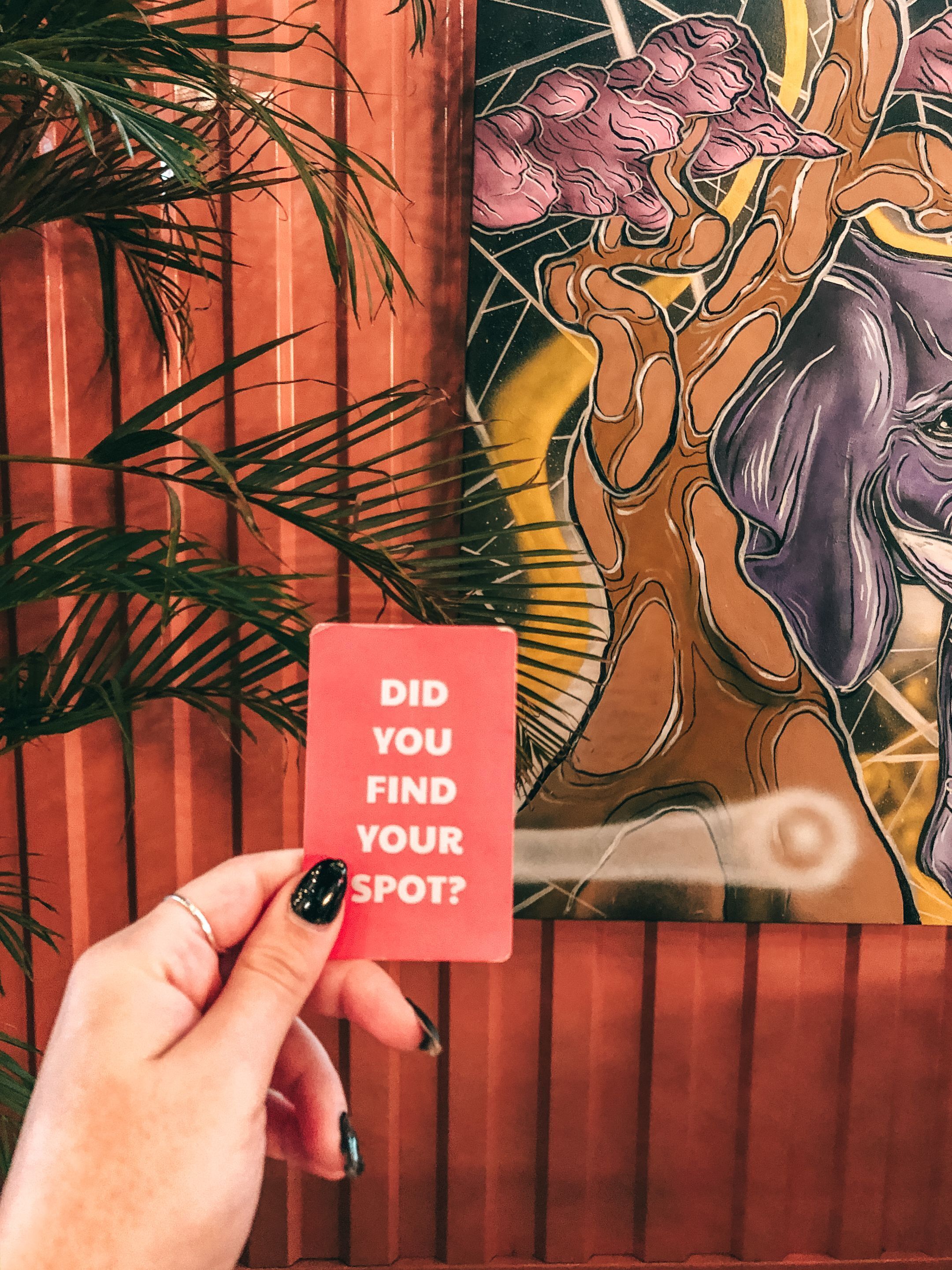 A hand holding a card with the text "DID YOU FIND YOUR SPOT?" in front of a colorful artwork.