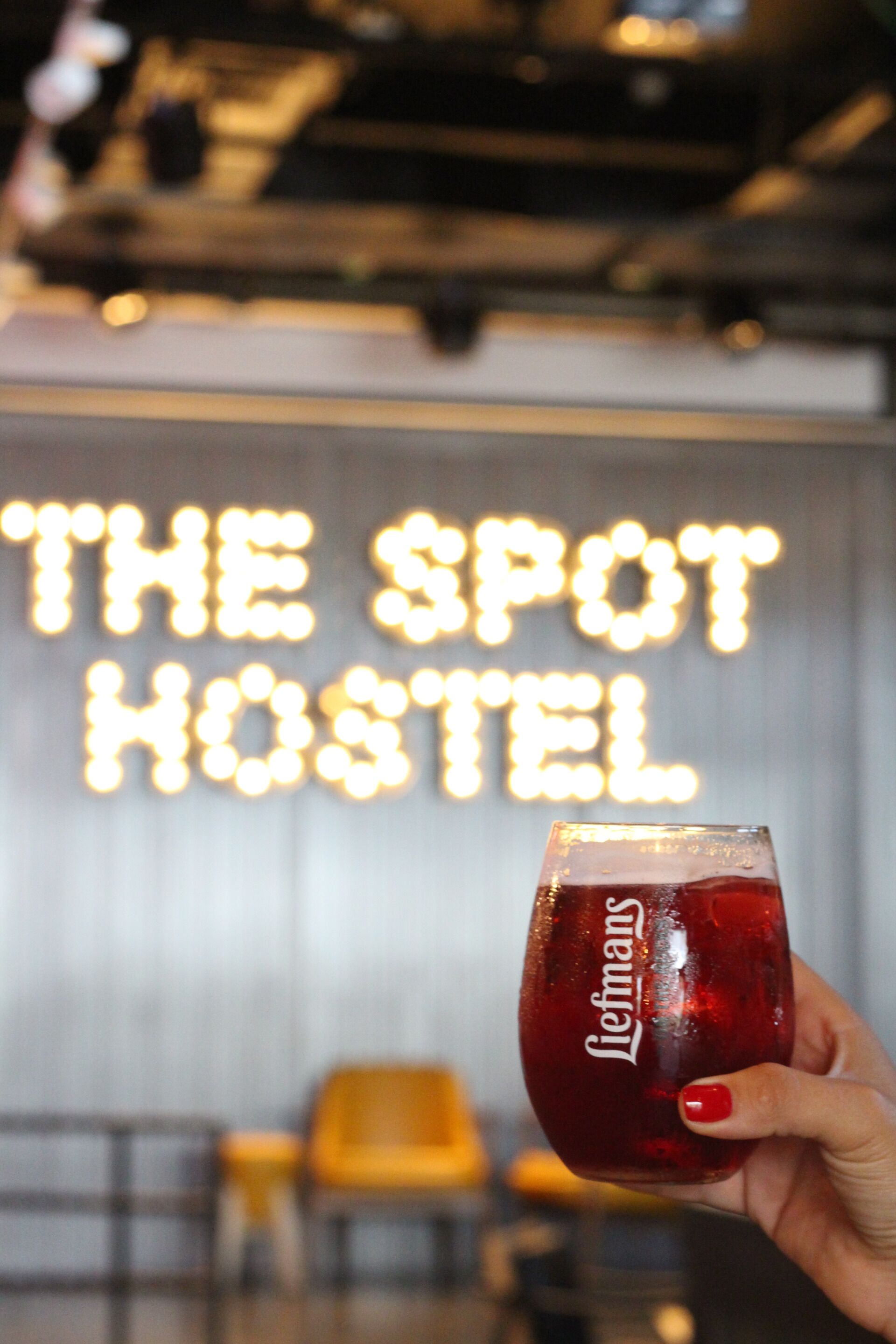 A hand holding a glass with a red liquid, in front of a neon sign that reads "THE SPOT HOSTEL".