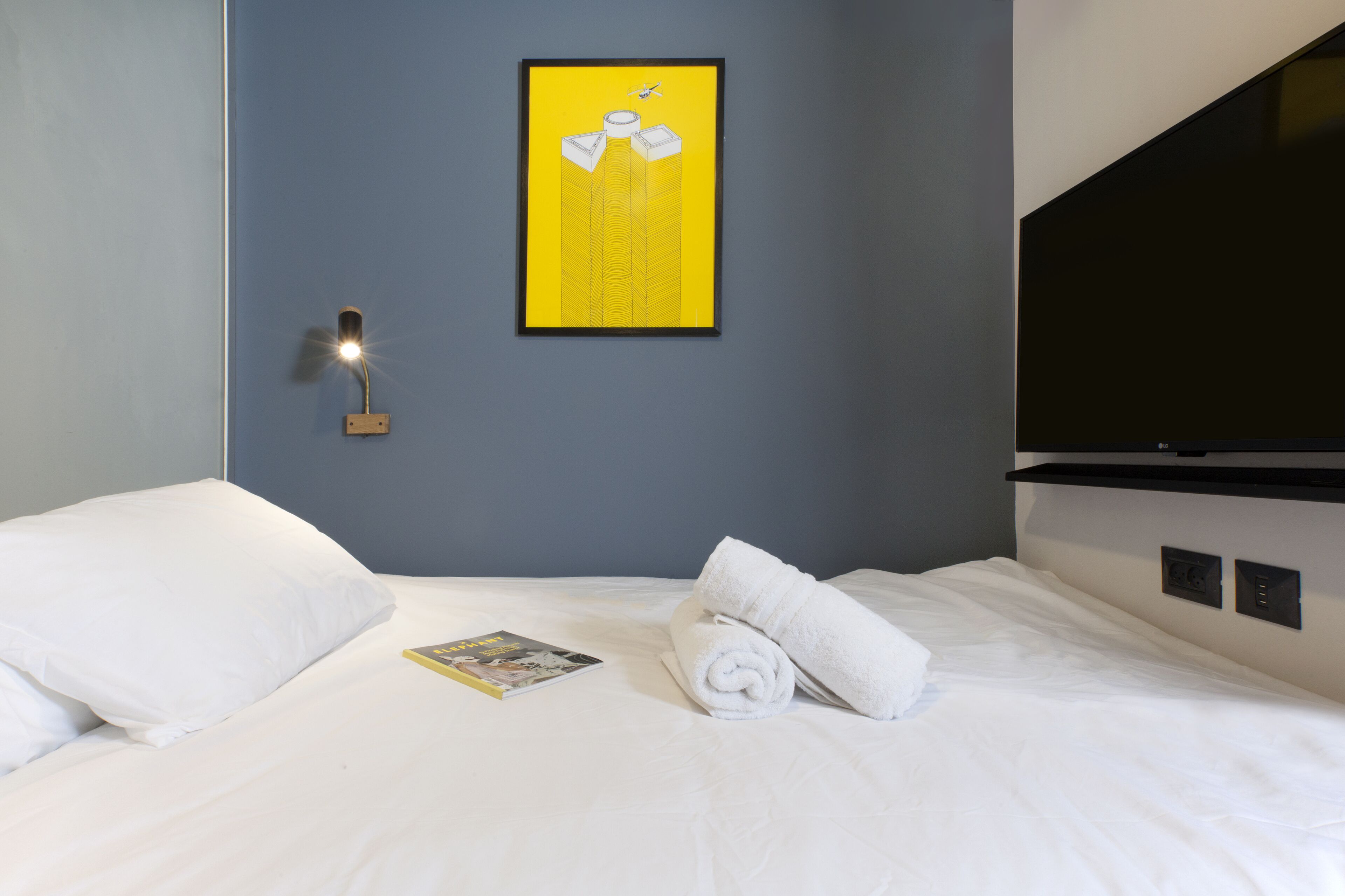 Ett hotellrum med en säng, en väggmonterad lampa, en inramad tavla och en hoprullad handduk på sängen.