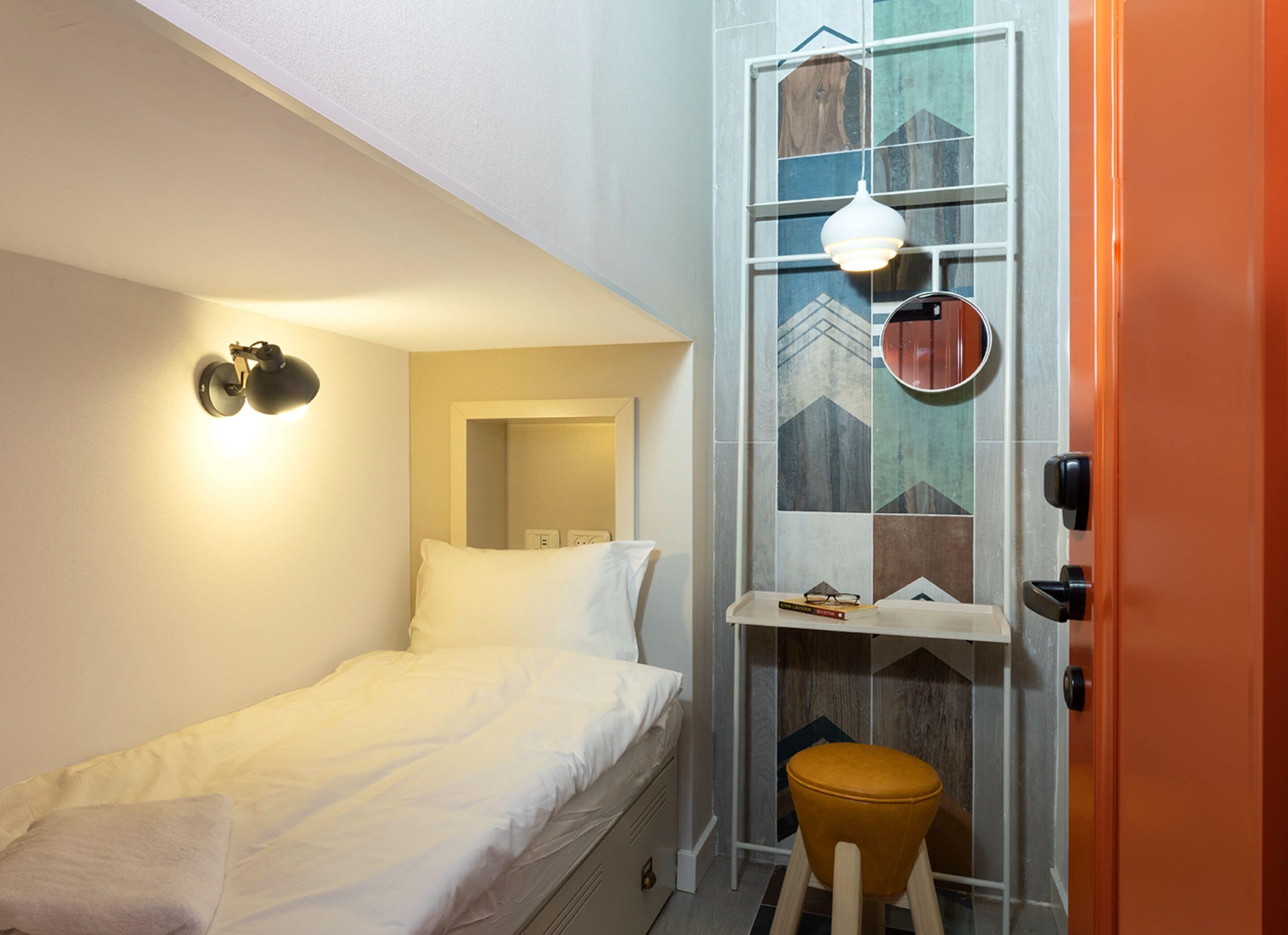 A small, compact room with a single bed, a stool, a shelf with books, and a wall-mounted light fixture.