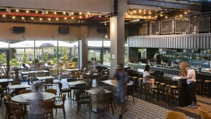 An indoor dining area with tables and chairs, a bar area with stools, and a view of the outdoors through large windows. - The Spot Hostel (Tel Aviv)