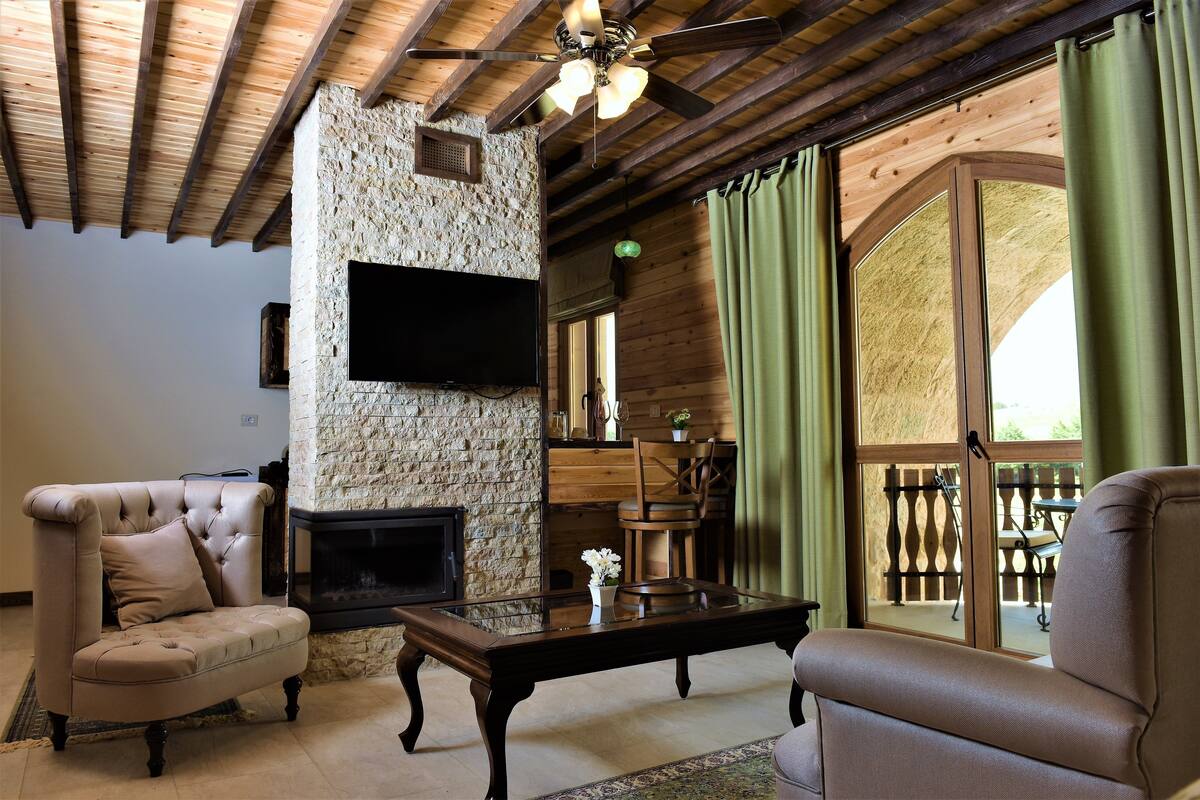 A cozy living room with a stone fireplace, wooden ceiling beams, and a large window with curtains.