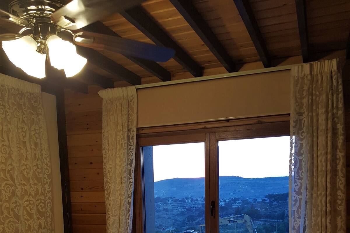 A bedroom with a wooden ceiling, a bed with a floral bedspread, a nightstand with a lamp, and a window with a view of a town and hills.