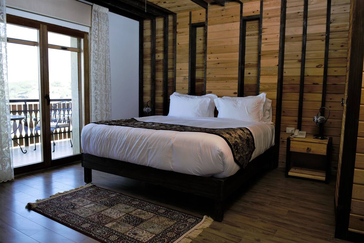 A bedroom with a large bed, wooden walls, a balcony with a railing, and a nightstand with a lamp.