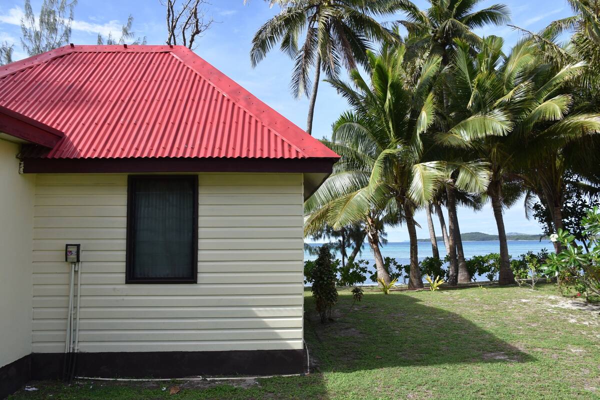 A small, single-story building with a red roof and a window. It is surrounded by a grassy area with palm trees and a view of the ocean.