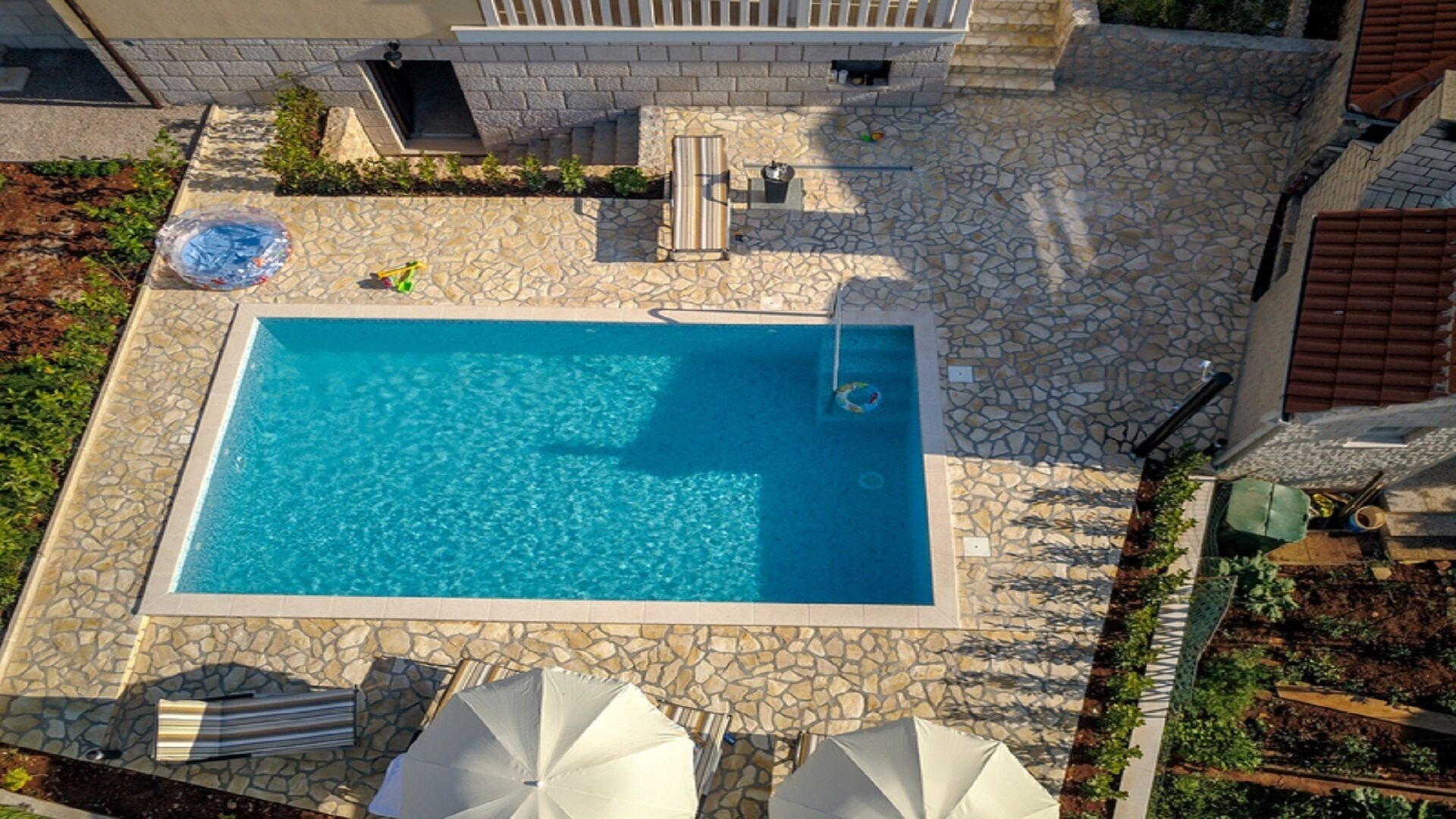 Une piscine rectangulaire avec une plage en pierre, entourée d’un mur de pierre et d’un petit jardin agrémenté de plantes et d’un banc.