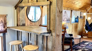 Un interior rústico con barra de madera, dos taburetes, un espejo redondo y una zona de estar acogedora.