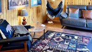 Interior acogedor de una cabaña con techo de madera, una silla hamaca, un sofá con cojines, un tapete estampado y decoración en la pared, incluyendo una imagen de un oso.