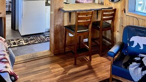 Interior acogedor de una cabaña con techo de madera, un comedor con dos sillas, un refrigerador y una cocina con lámpara colgante.