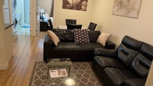 Ein Wohnzimmer mit einer schwarzen Ledersofa, einem Glastisch und einem gemusterten Teppich. An der Wand hängt ein Gemälde, und eine Tür führt ins Badezimmer.