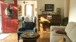 A living room with a red door, a blue armchair, a wooden coffee table, a beige sofa, and a piano.