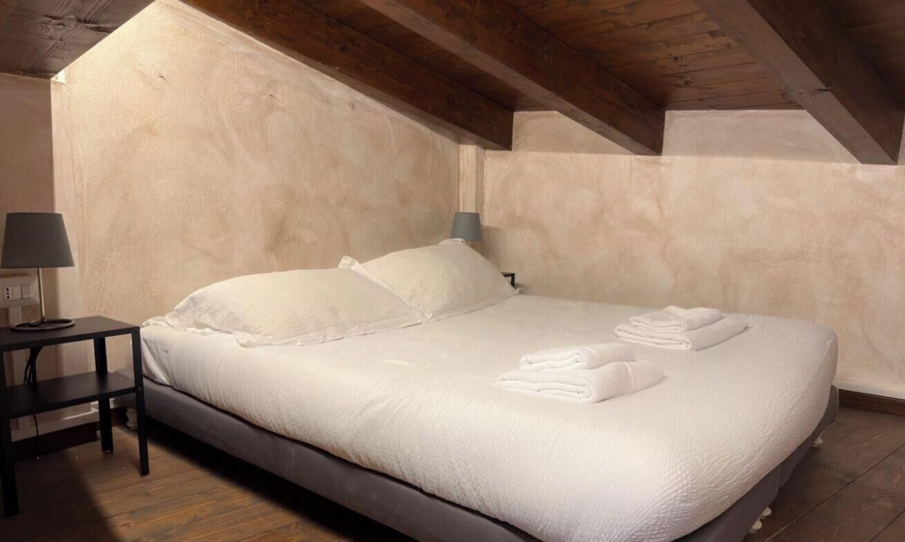 A single bed with white linens and pillows, a bedside table with a lamp, and a wooden ceiling.
