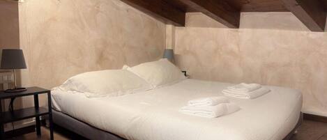 A single bed with white linens and pillows, a bedside table with a lamp, and a wooden ceiling.