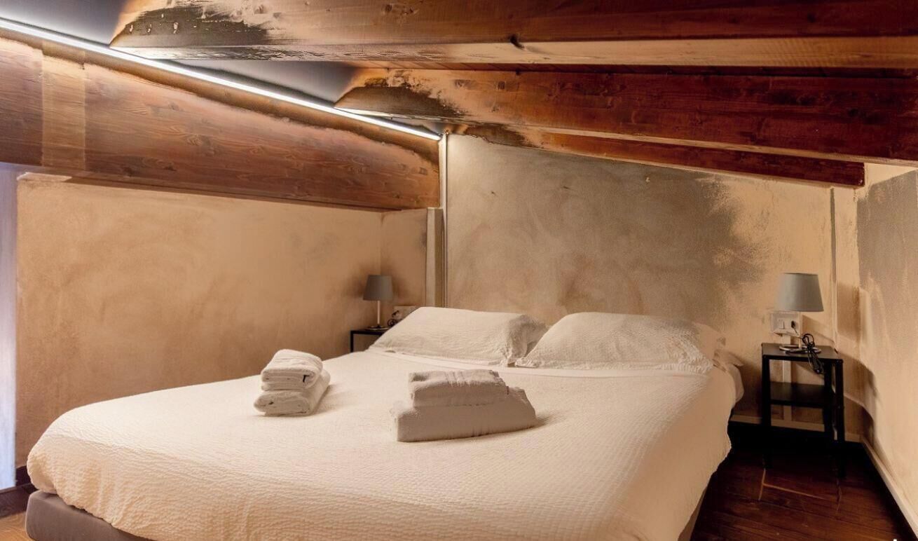 Una cama individual con una colcha blanca y dos almohadas, una mesita de noche con una lámpara, y una pared con vigas de madera a la vista.