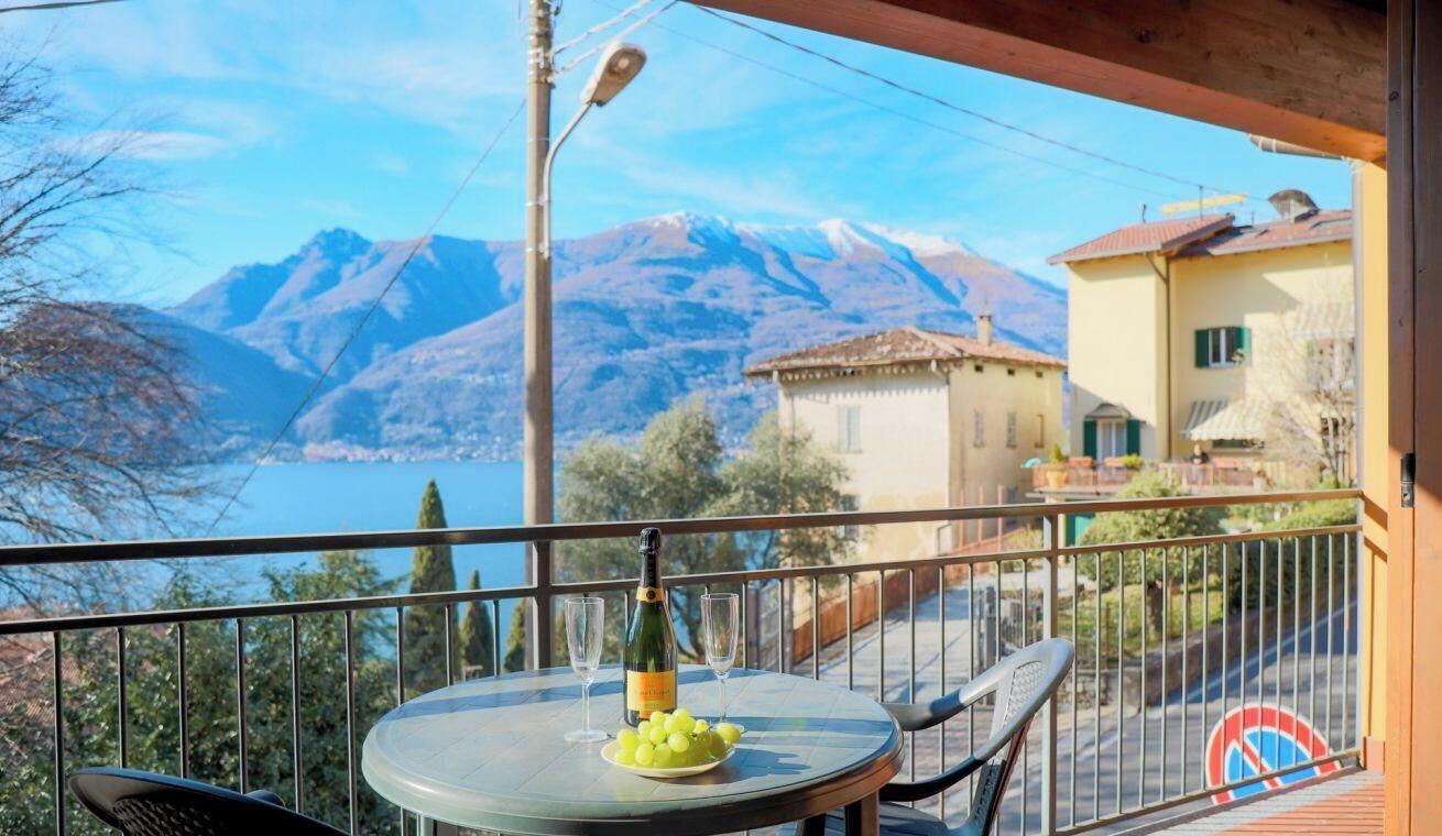 Un balcón con una mesa, una botella y vasos, con vistas a un lago y montañas.