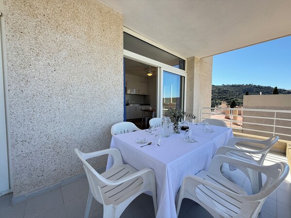 A balcony with a table set for dining, white plastic chairs, and a view of the surrounding area.