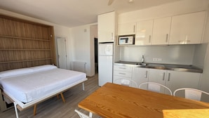 Een kleine kamer met een bed, een houten tafel, een kitchenette en een witte radiator.