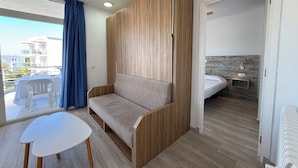 Een moderne kamer met een bank, een klein tafeltje, een bed met houten hoofdeinde en een balkon met uitzicht op gebouwen.