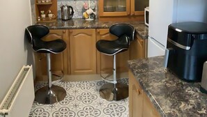 Une cuisine avec deux tabourets de bar noirs, un panneau indiquant « café et thé », des placards en bois et un four à micro-ondes.