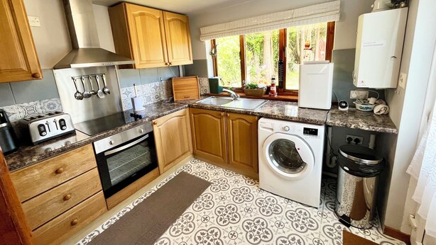 Une cuisine compacte avec des meubles en bois, un four encastré, un lave-linge et une petite fenêtre donnant sur un espace verdoyant.