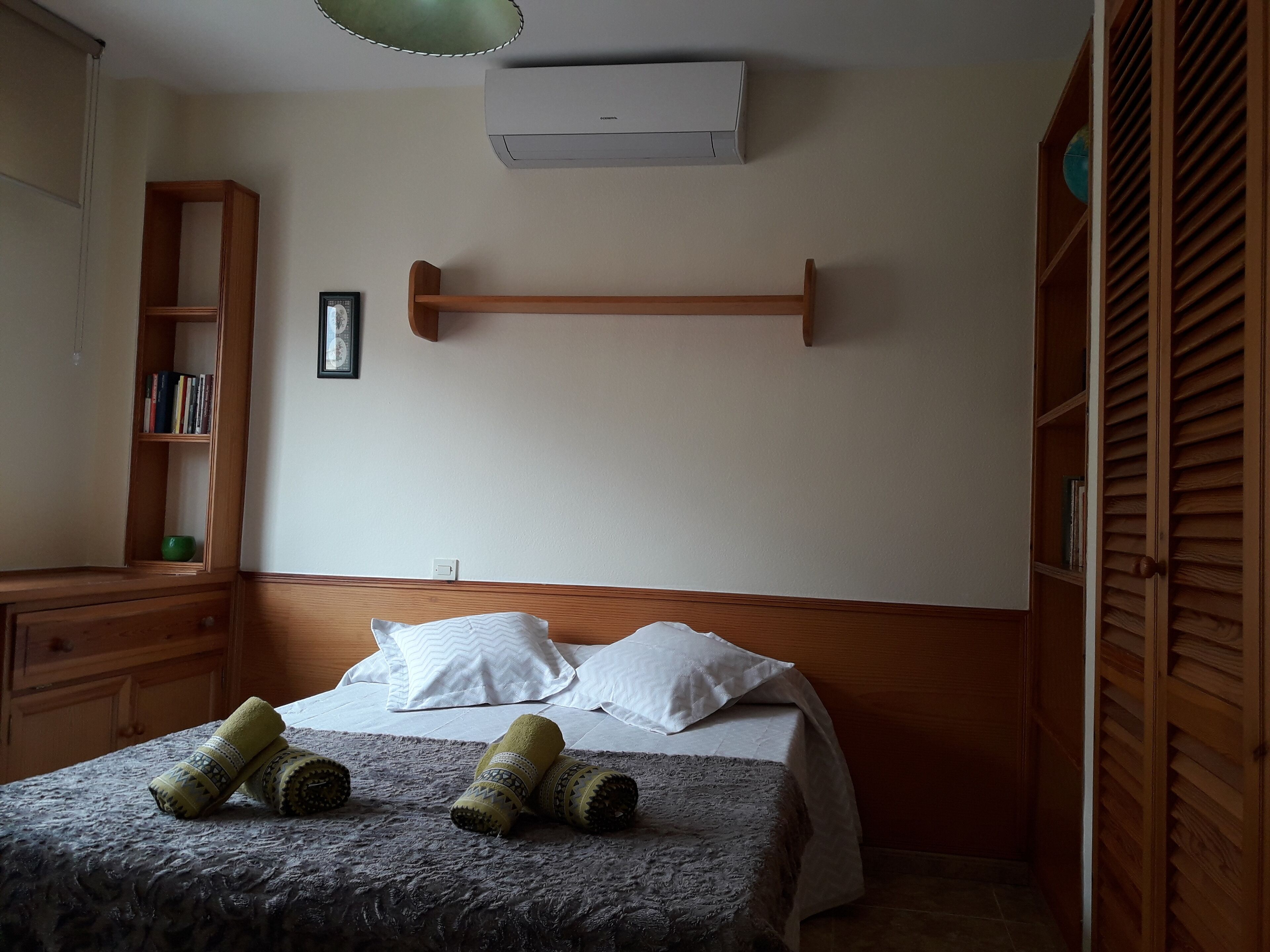 Un dormitorio con cama, cabecera de madera, mesita de noche y estantería, además de un equipo de aire acondicionado.
