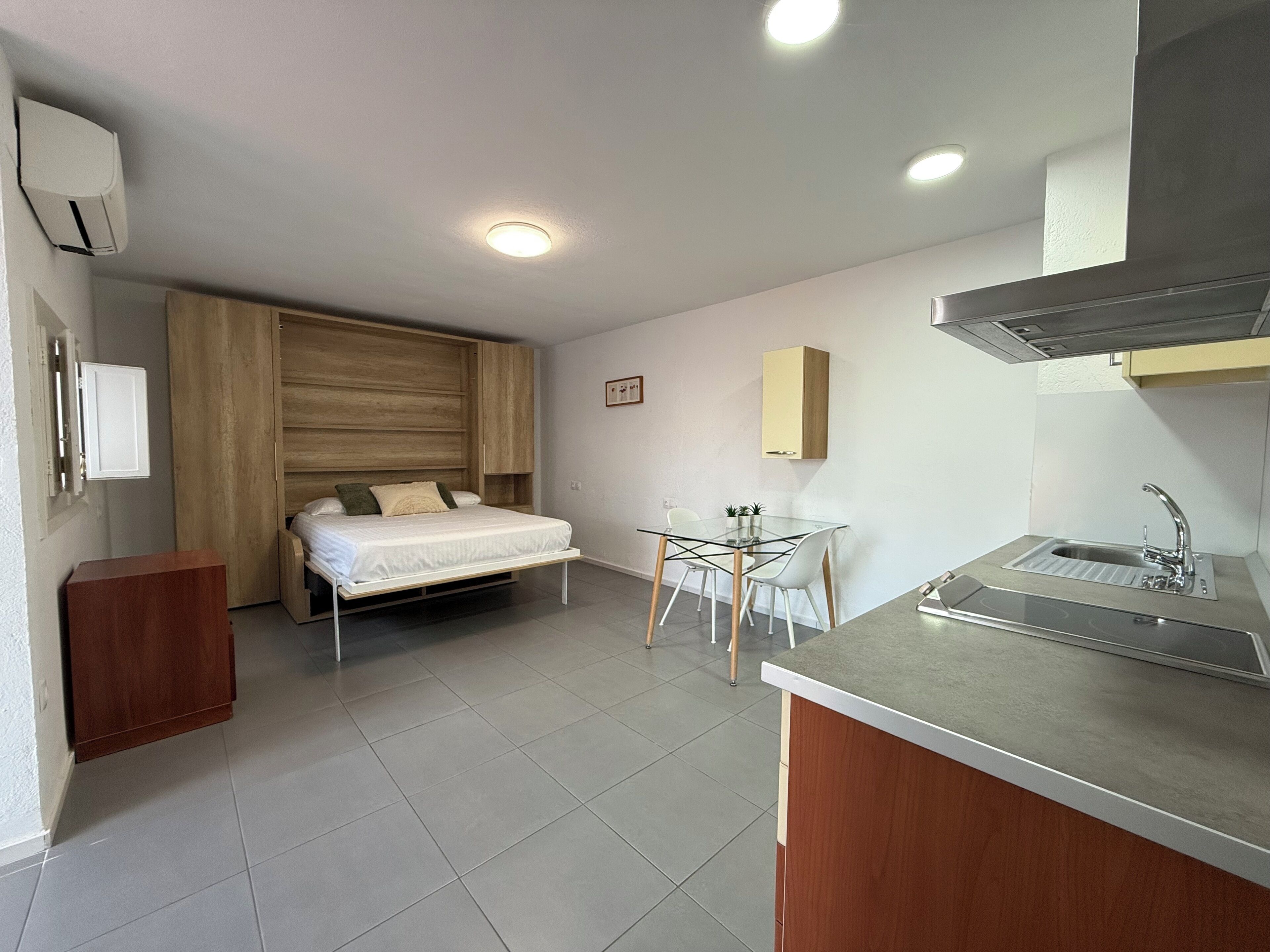 A compact bedroom with a bed, bedside table, and a small desk with a chair. There is a kitchenette with a sink and a stove.