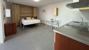 A compact bedroom with a bed, bedside table, and a small desk with a chair. There is a kitchenette with a sink and a stove. - Agi Peater Studio Apartamento for rent in Roses Girona. (Rosas / Roses)