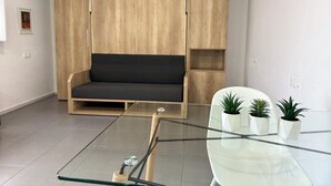 A modern living room with a wooden wardrobe, a dark sofa, a glass coffee table, and three potted plants. - Agi Peater Studio Apartamento for rent in Roses Girona. (Rosas / Roses)