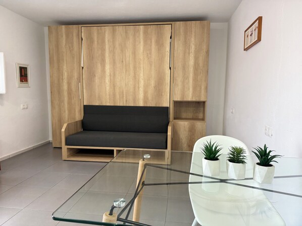 A modern living room with a wooden wardrobe, a dark sofa, a glass coffee table, and three potted plants. - Agi Peater Studio Apartamento for rent in Roses Girona. (Rosas / Roses)