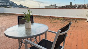 A rooftop terrace with a round glass table and two chairs, a potted plant, and a view of buildings and hills in the background. - Agi Peater Studio Apartamento for rent in Roses Girona. (Rosas / Roses)