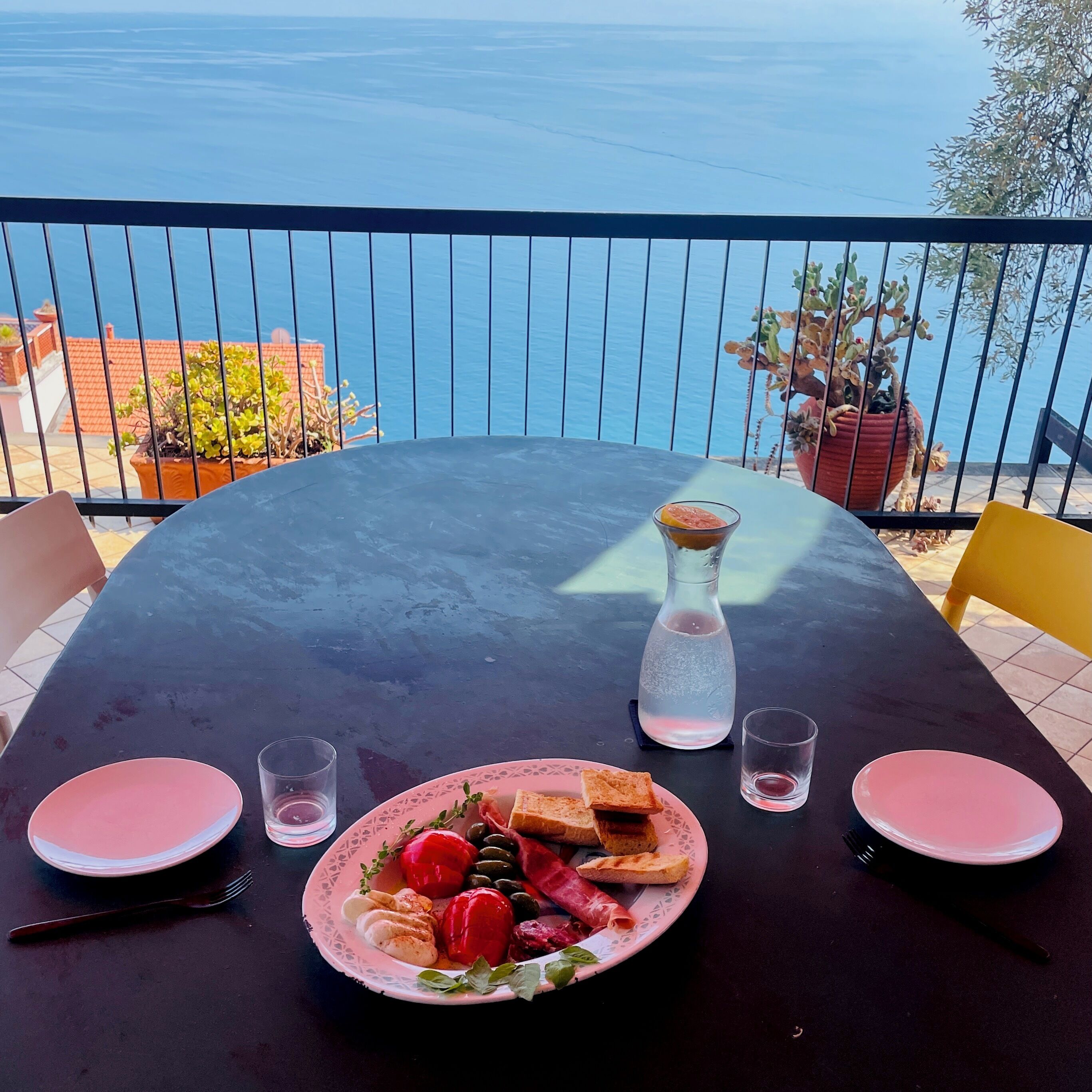 A table set for two with a meal, a bottle of water, and glasses on a balcony overlooking the sea.
