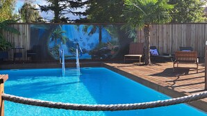 En swimmingpool med træterrasse, omgivet af et bambushegn og et tropisk muralmaleri.
