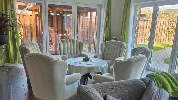 En stue med et rundt bord, fire lænestole og en sofa. Der er en skydedør, som fører til et udendørs område med en træterrasse og stole.