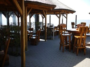 Una terraza de madera al aire libre con mesas y sillas, techo de paja y vista al mar.