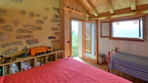 Ein gemütliches Zimmer mit Steinwand, einem Bett mit roter Steudecke, einer Holzbank und einem Fenster mit Blick auf Grünflächen.